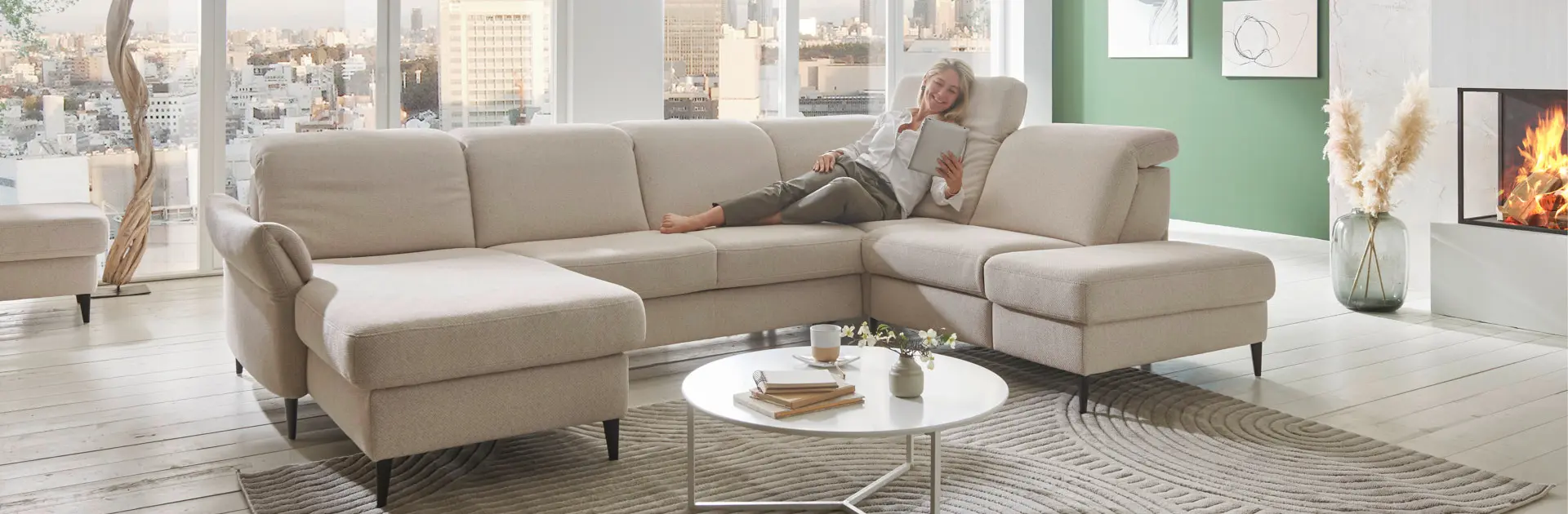 Helles Wohnzimmer mit großem beigem Ecksofa, entspannter Frau mit Tablet, Kaminfeuer und Panoramablick über die Stadt